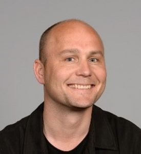 Man with a big smile standing against a neutral background.