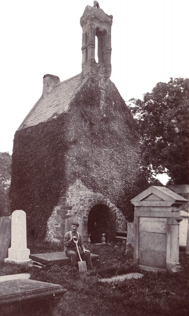 A photography from the 19th century showing a Scottish sexton with a spade siting in a kirkyard next to a small kirk.