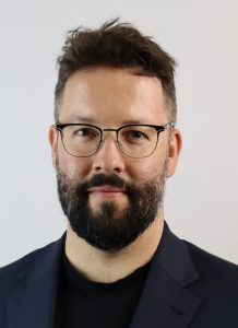 Man with glasses and a beard looks into camera.